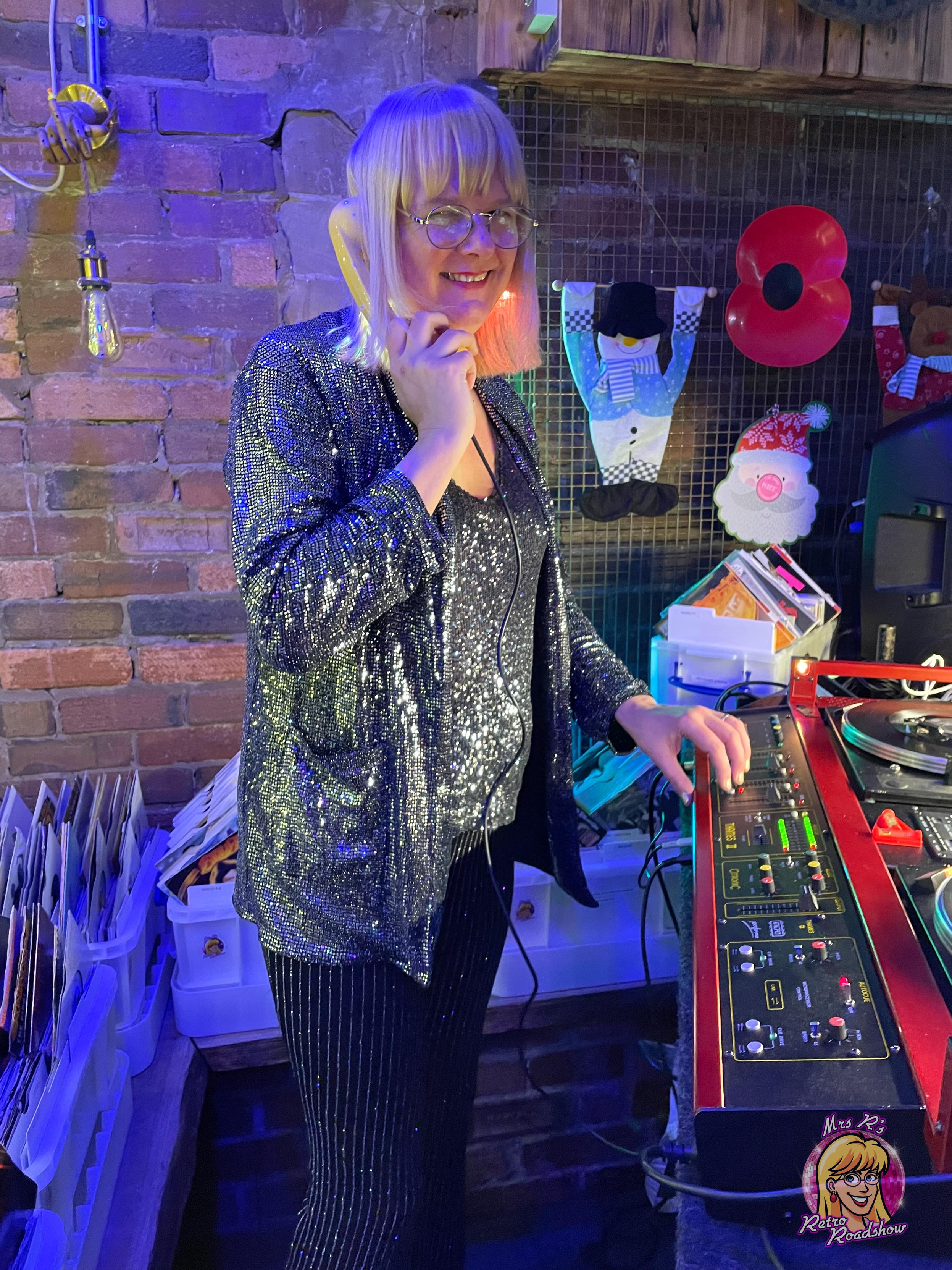 A blonde woman stands at a 1980s disco console, smiling at the camera and with one hand on the console. She is holding a vintage phone handset to her ear. Behind her are 7" vinyl records.