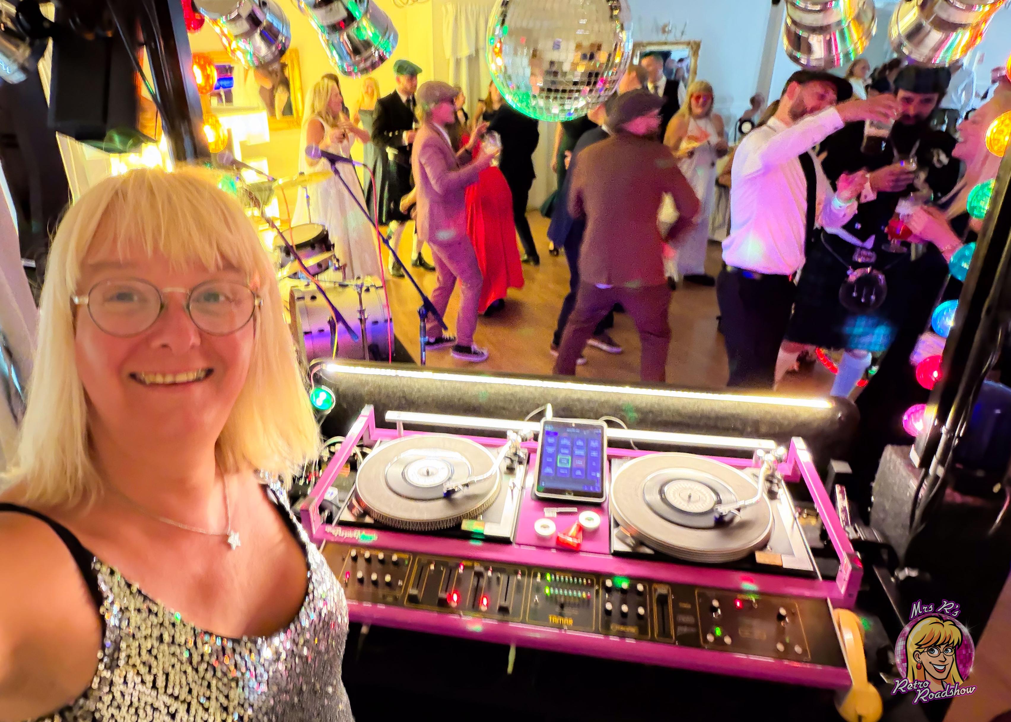 Selfie of a blonde woman smiling at the camera, she's wearing sequins and standing at a 1980s disco console playing 7" vinyl records. In the background, people in smart clothes and wedding attire are dancing.